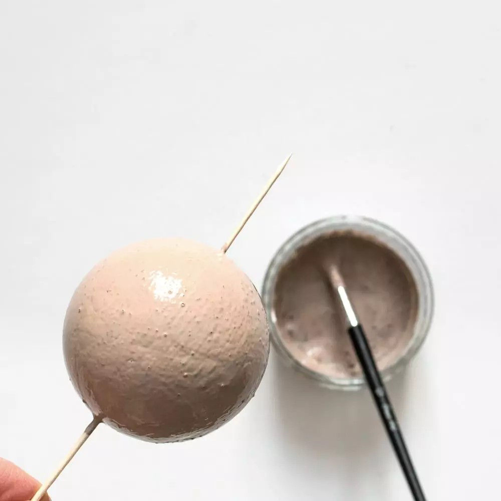 Une sphere texturee est maintenue sur une brochette en bois, placee pres d'un pot contenant de la peinture ou du materiel de bricolage. Un pinceau repose a l'interieur du pot, suggerant une activite artistique ou de bricolage. La palette neutre met l'accent sur une esthetique creative et minimaliste.