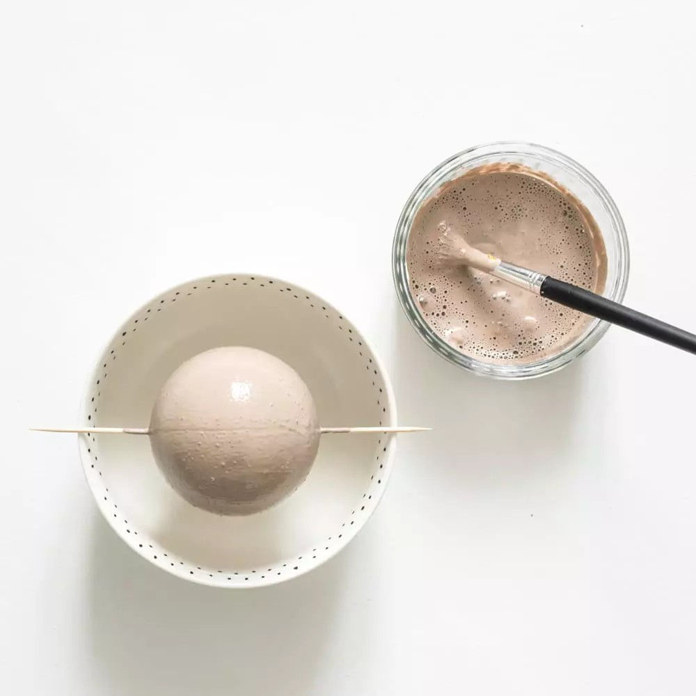 Un objet spherique est suspendu au-dessus d'un bol blanc, maintenu en place par un b�ton en bois. Un recipient en verre rempli de peinture beige et un pinceau sont places a proximite, suggerant une activite manuelle. Le fond blanc immacule souligne la simplicite et met l'accent sur les materiaux.