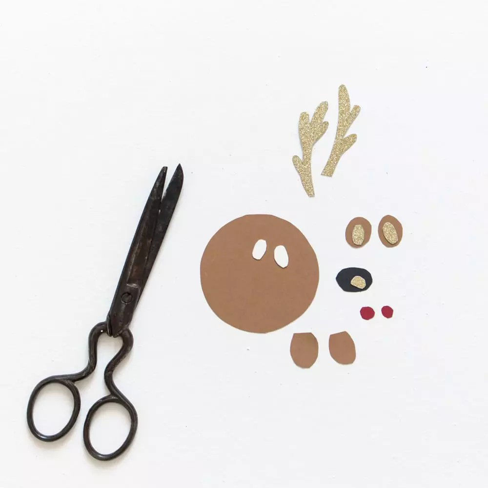 Une paire de ciseaux de style vintage est posee a c�te de decoupes en papier formant un visage de renne. Les decoupes comprennent des bois, des yeux, un nez et des oreilles dans des tons ocres avec des accents scintillants. Le fond blanc minimaliste souligne le theme du bricolage.