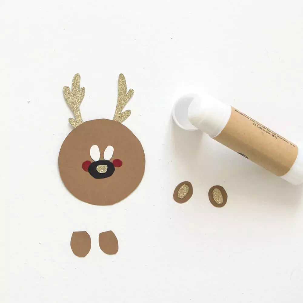 Des decoupes pour un bricolage representant un renne, notamment un visage circulaire marron, des bois scintillants et de petites oreilles, sont disposees sur une surface blanche. Un b�ton de colle avec une etiquette marron est place a proximite, suggerant une activite de bricolage. La scene est minimaliste et met l'accent sur la creativite et le bricolage.