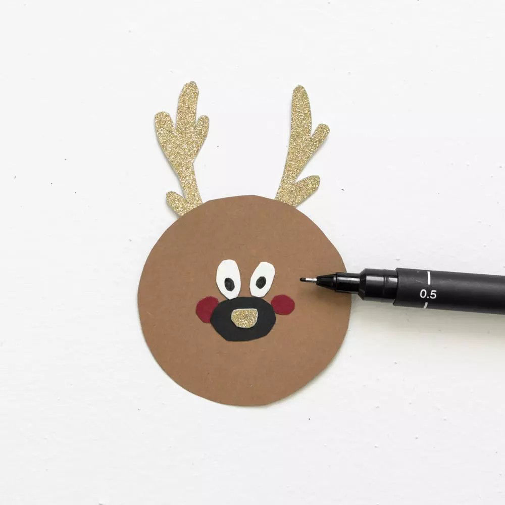 Un renne circulaire fabrique a partir de papier brun avec des bois dores scintillants et des traits du visage peints. Le dessin comprend des yeux blancs, un nez noir et des joues rouges, avec un stylo noir marque � 0,5 � ajoutant des details. Le bricolage est place sur un fond blanc uni, soulignant sa simplicite et son charme.
