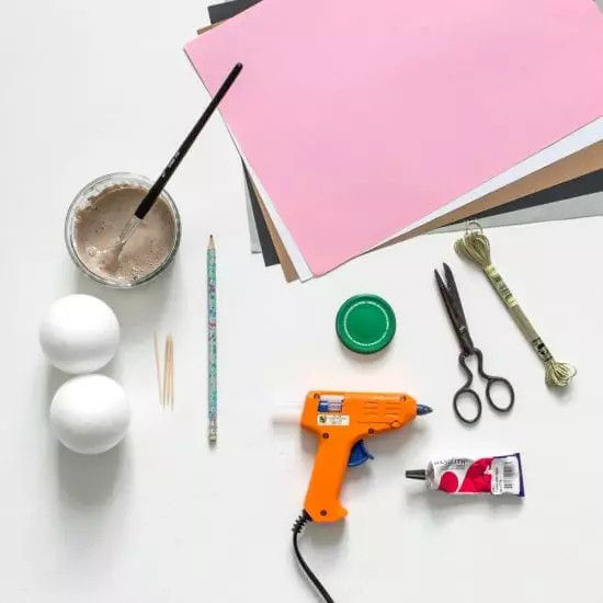 Une collection de fournitures de bricolage disposees sur une table blanche, comprenant un pistolet a colle chaude, des ciseaux, un pinceau et du papier colore. Deux boules en mousse blanche, un bouchon de bouteille vert et un tube de peinture rouge sont egalement visibles. Parmi les autres articles, on trouve des cure-dents, un crayon et un metre ruban.
