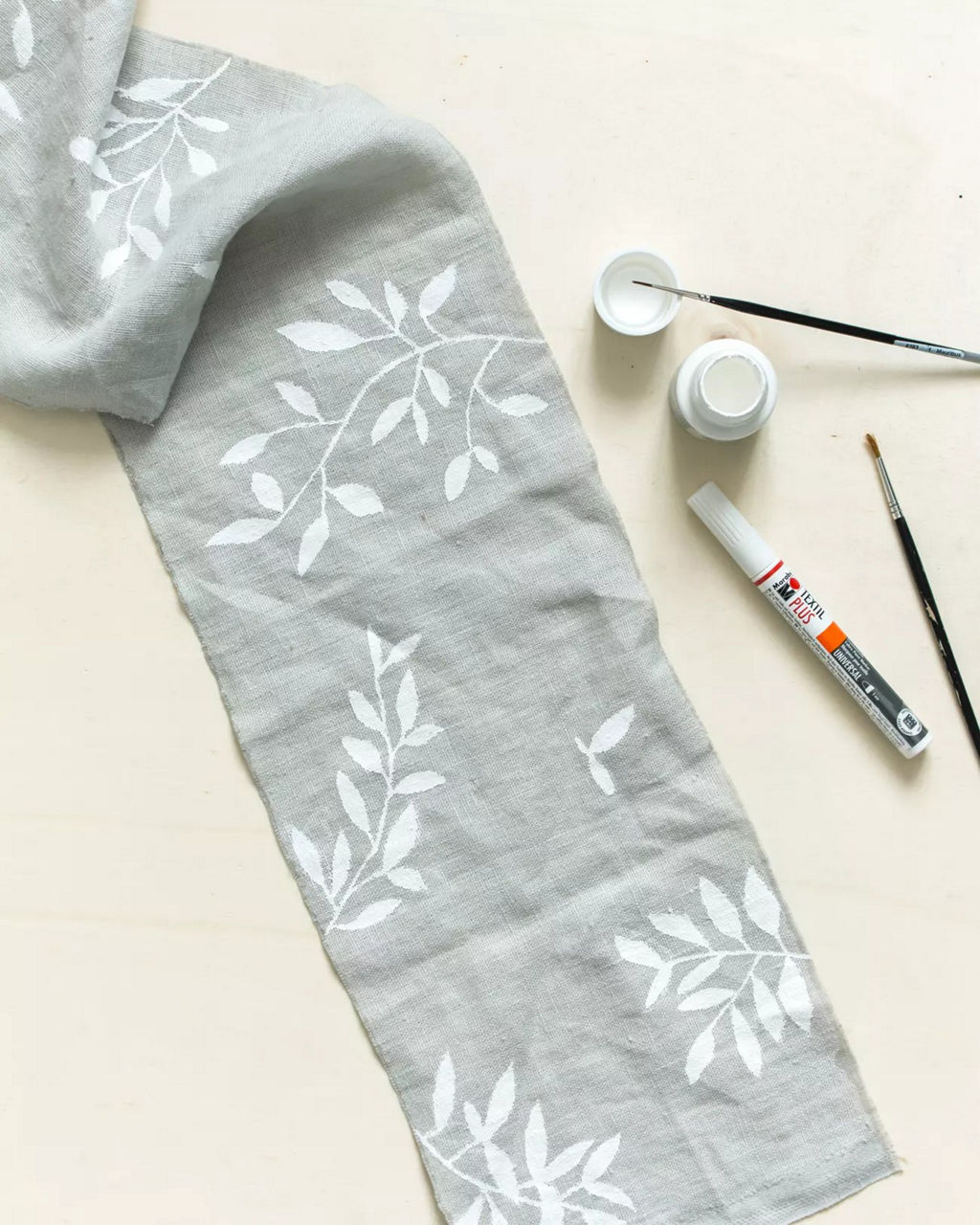 Un tissu gris clair orne de motifs floraux blancs peints a la main est presente a c�te de fournitures artistiques. Le decor comprend des pinceaux, un marqueur a peinture et des pots de peinture ouverts, suggerant une activite creative. Le motif presente de delicats motifs de feuilles, creant une esthetique minimaliste et elegante.