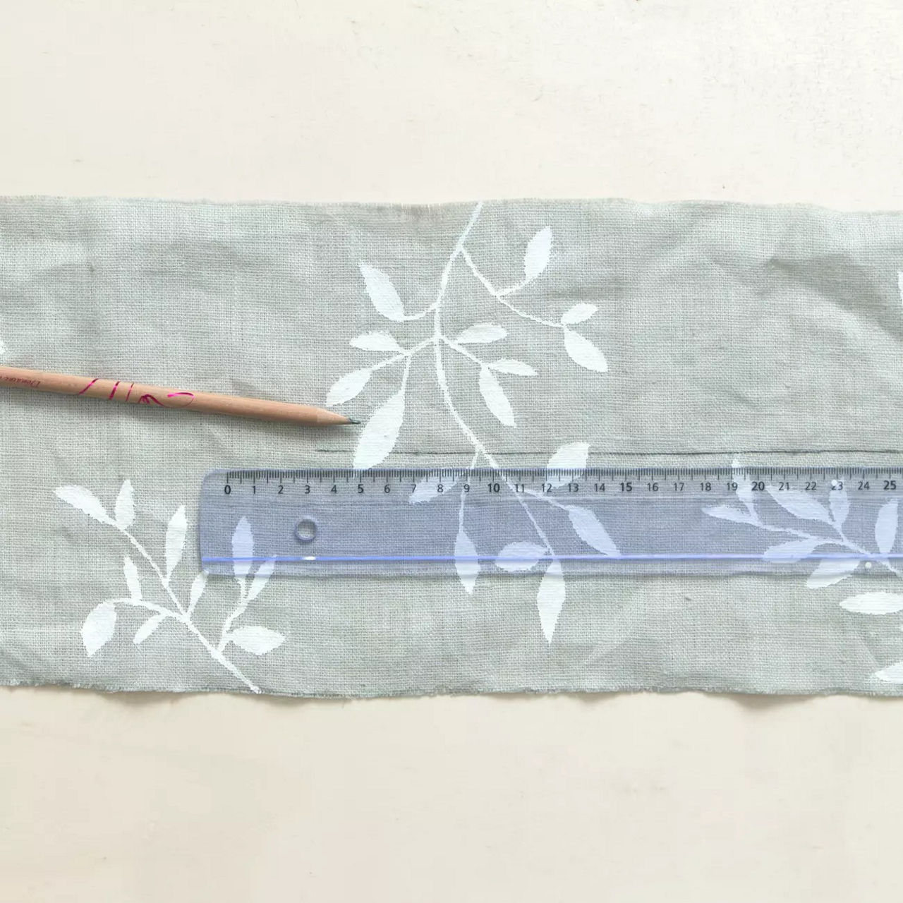 Un tissu a motifs de feuilles blanches est presente a c�te d'une regle transparente et d'un b�ton en bois. La regle indique les mesures en centimetres, avec des reperes clairement visibles jusqu'a 30 cm. Le decor est minimaliste et neutre, mettant en valeur la texture et le motif du tissu.
