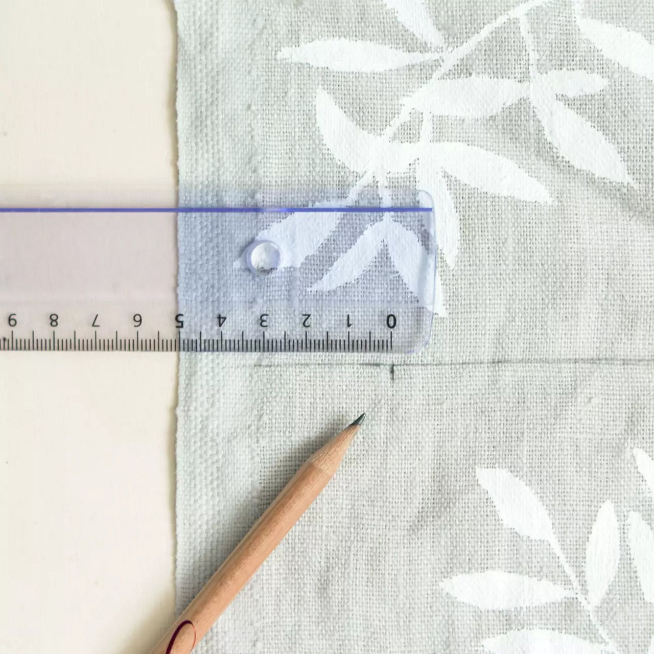 Une regle transparente et un crayon en bois sont places sur un tissu de couleur claire avec un motif floral blanc. La regle affiche des mesures metriques claires, avec des chiffres allant jusqu'a 15 cm. Le decor suggere des activites de bricolage ou de couture, avec des textures douces et des tons neutres creant une ambiance calme et creative.