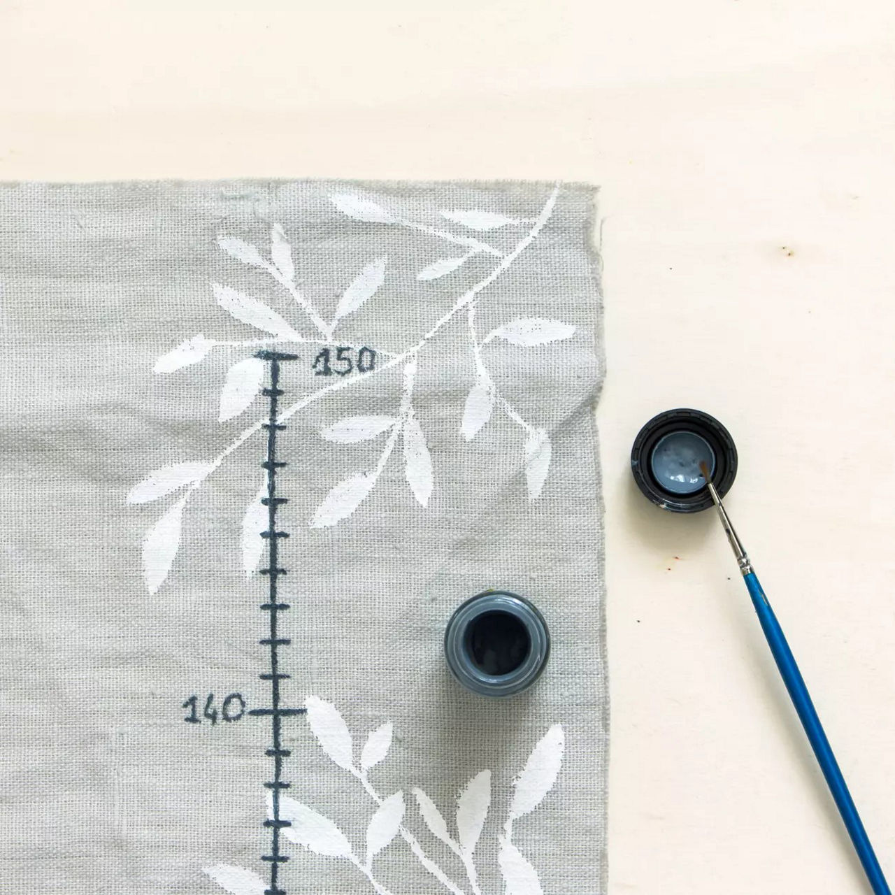 Gros plan sur un tissu peint a la main presentant de delicats motifs floraux blancs sur fond gris. Le motif comprend des marques numeriques visibles, telles que � 140 � et � 150 �, ressemblant a une echelle de mesure. Un petit pinceau et un pot ouvert de peinture bleu fonce sont places a proximite, suggerant un decor artistique. L'image traduit un processus de creation creatif et detaille.
