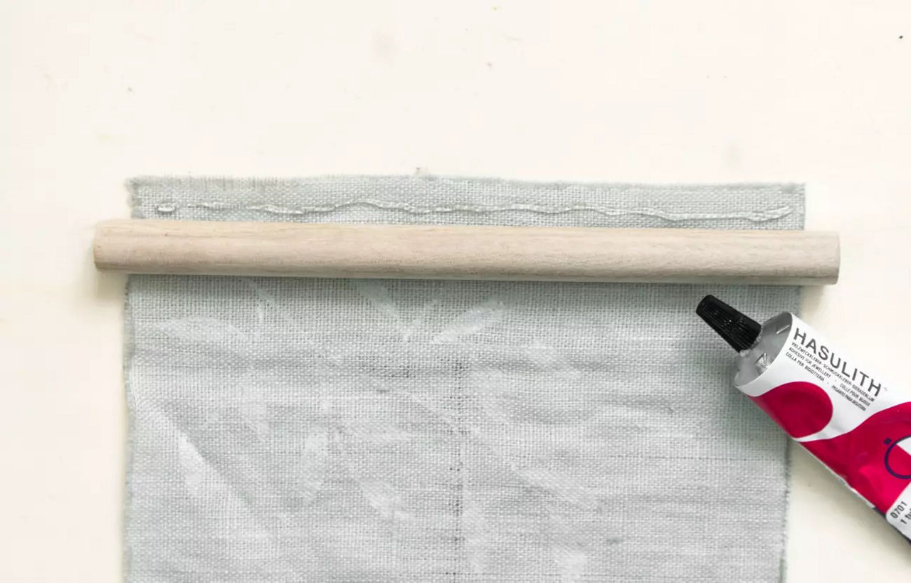 Un morceau de tissu gris clair est partiellement enroule sur un goujon en bois, accompagne d'un tube de colle portant l'inscription � MASULITH � avec un motif rouge et blanc. Le decor est minimaliste et epure, mettant l'accent sur le processus de fabrication ou d'assemblage. Le tube de colle est debouche, ce qui suggere une utilisation active.