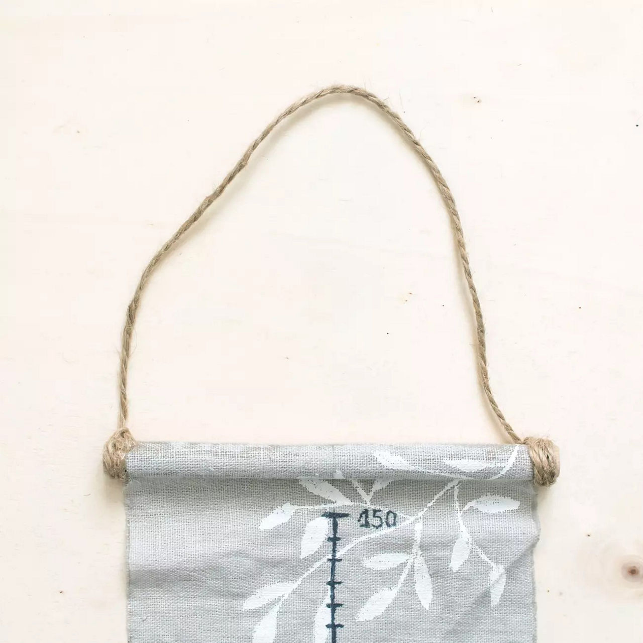 Un tableau de croissance en tissu presentant un motif minimaliste de feuilles imprime sur un fond beige neutre. Le tableau est marque de mesures visibles, notamment � 150 �, et est suspendu a une boucle en corde naturelle. Le motif est simple et rustique, adapte a la decoration interieure ou aux chambres d'enfants. La palette de couleurs douces s'harmonise avec une variete de styles d'interieur.