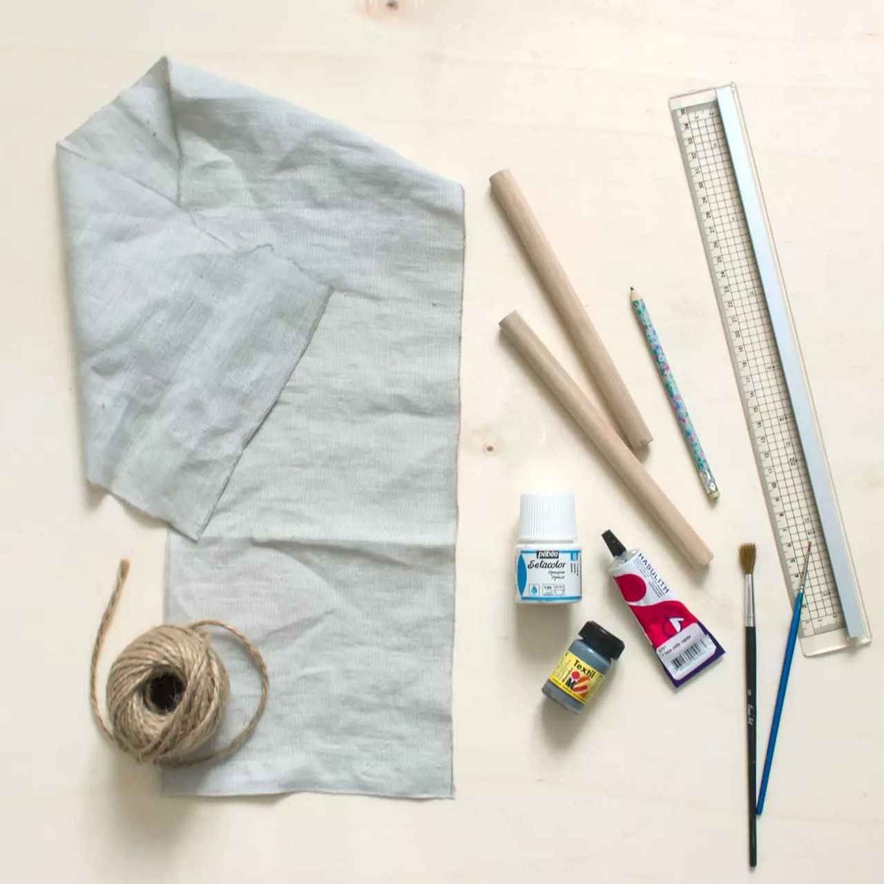 Une collection de materiaux de bricolage comprenant un morceau de tissu, de la ficelle, des chevilles en bois et divers outils. La composition comprend une regle, un crayon, un pinceau et des produits adhesifs, disposes de maniere ordonnee sur une surface en bois clair. Les articles visibles comprennent un flacon de colle de 50 ml et un tube de colle rouge.