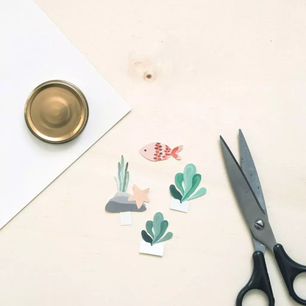 Une composition a plat de materiaux de bricolage comprenant des ciseaux, un objet circulaire dore et des decoupes de papier sur le theme aquatique, notamment un poisson, une etoile de mer et des plantes marines. Le decor est une surface en bois clair avec une decoration minimaliste. La scene evoque la creativite et les activites de bricolage.