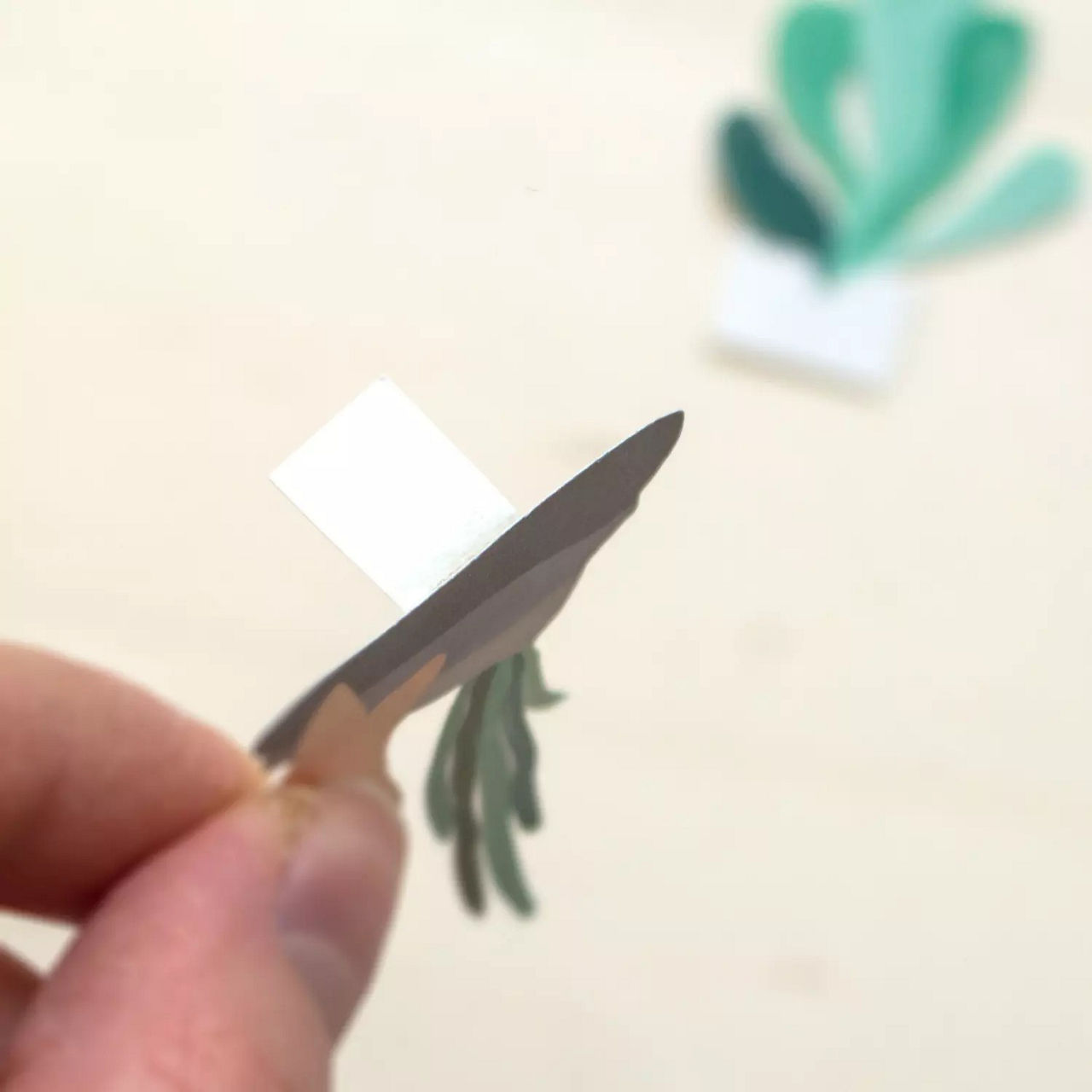 Gros plan sur une main tenant une petite decoupe en papier ressemblant a une plante. L'arriere-plan presente des formes floues vertes et blanches, suggerant un cadre creatif ou artisanal. L'image met l'accent sur la precision et les details de l'art du papier.