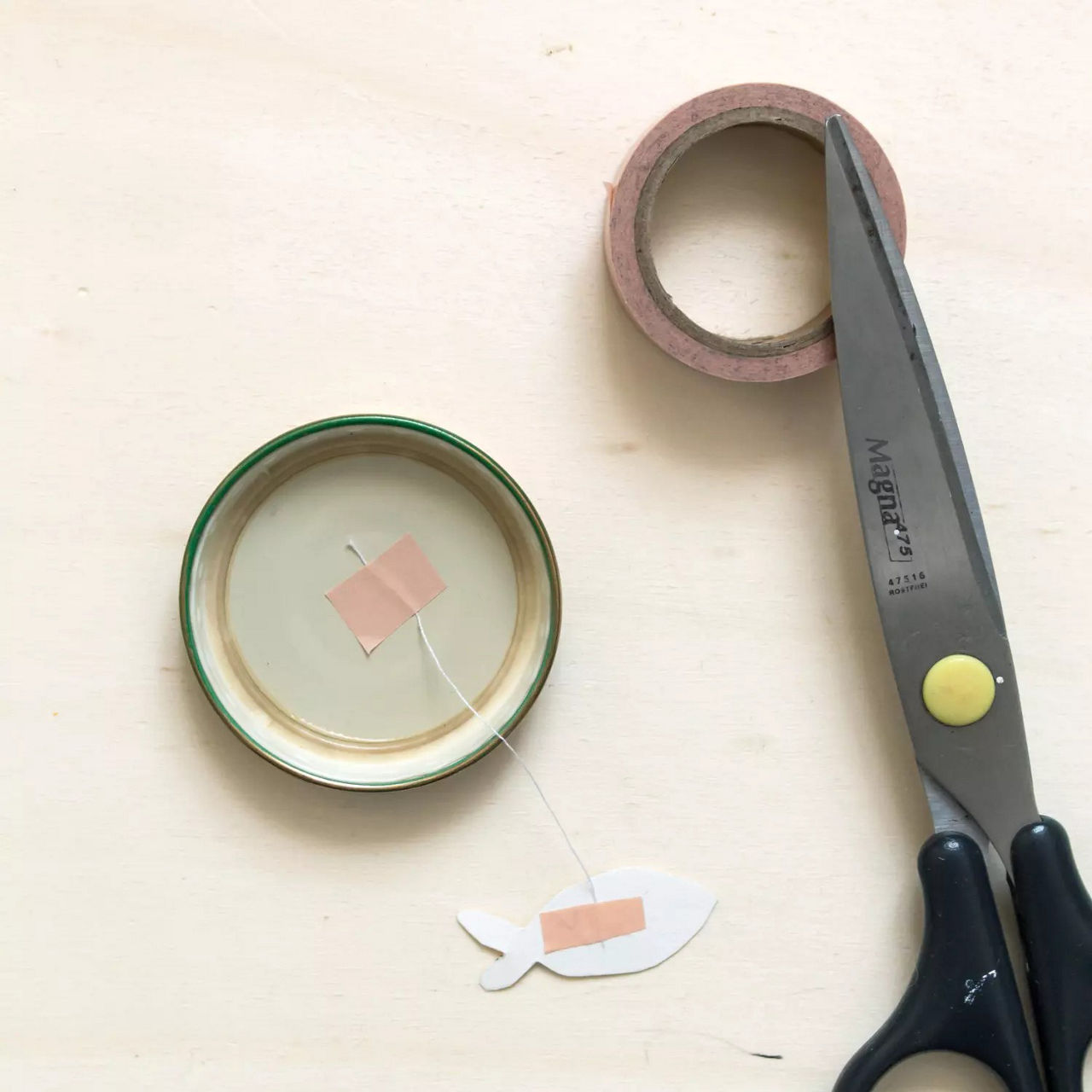 Une paire de ciseaux portant l'inscription � Magnet 21 cm � est posee sur une surface en bois clair a c�te d'un projet de bricolage. Le projet comprend une decoupe de poisson attachee a une ficelle et un couvercle de bocal avec du ruban adhesif. Un rouleau de ruban adhesif rose est egalement visible, suggerant une activite creative d' . Le decor est minimaliste et organise, mettant l'accent sur les outils et les materiaux utilises.