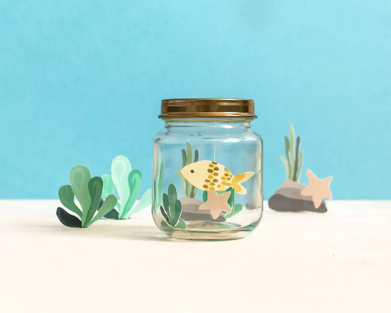 Un petit bocal en verre avec un couvercle dore contient un poisson jaune decoratif et des elements sur le theme aquatique. Le bocal est entoure de plantes marines et d'etoiles de mer en papier, sur un fond bleu vif. La scene evoque une esthetique sous-marine ludique et creative.