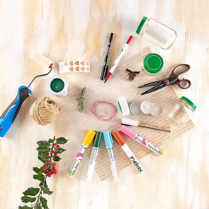 Une collection d'outils et de fournitures de bricolage disposes sur une surface en bois clair. Les articles comprennent des marqueurs colores, des ciseaux, de la ficelle, un rouleau a peinture et un bocal en verre avec un couvercle vert. Des elements decoratifs tels que des feuilles de houx et des bougies ajoutent une touche festive. Le texte visible comprend � Marabu � sur les marqueurs.