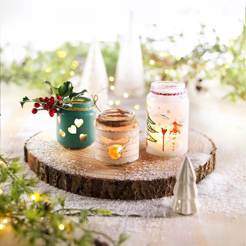 Trois pots a bougies decoratifs sont exposes sur une planche de bois rustique entouree d'elements sur le theme des fetes. Chaque pot presente des motifs uniques, notamment des decoupes en forme de c�ur, un emballage en ecorce de bouleau et des motifs hivernaux peints tels que des bonhommes de neige et des arbres. Le decor comprend des guirlandes lumineuses, des textures ressemblant a de la neige et de petites decorations festives, creant une ambiance chaleureuse pour les fetes.