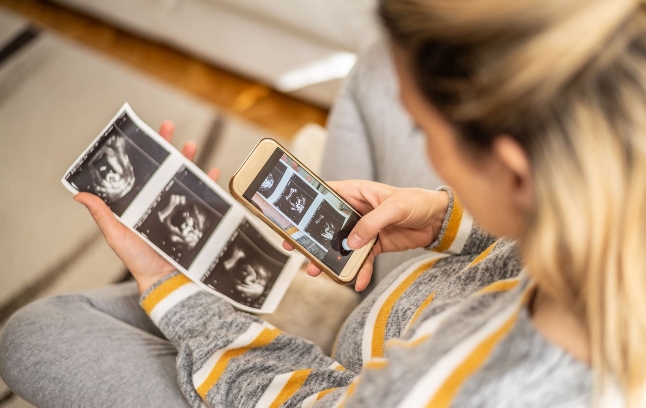 Une femme tient des images echographiques imprimees dans une main tout en utilisant un smartphone pour visualiser les versions numeriques. Elle est assise dans un cadre interieur confortable, vetue d une tenue decontractee grise et jaune. La scene met en evidence un moment personnel de reflexion et d utilisation de la technologie.