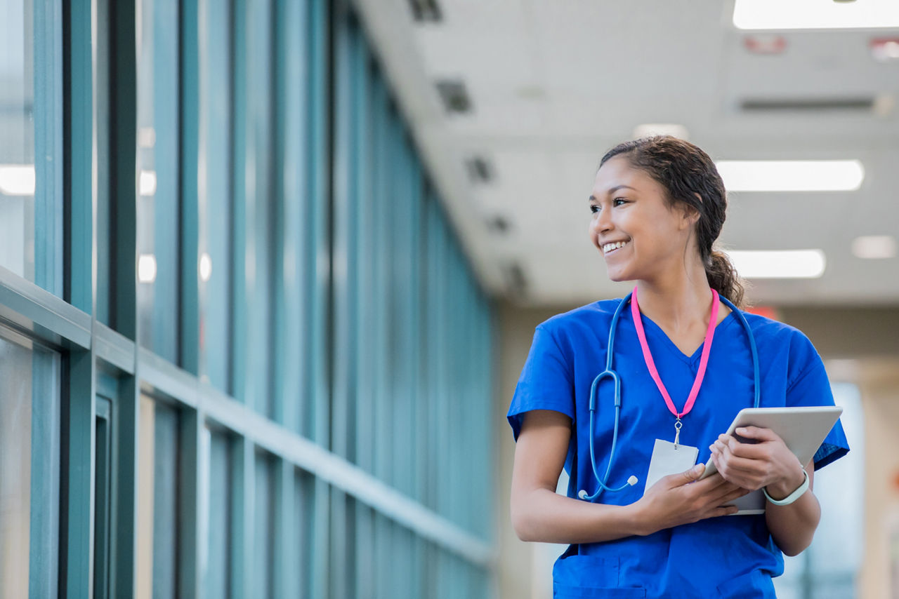 Happy young adult mixed race woman is nurse or healthcare worker. She is carrying a digital tablet patient chart and looking out the window of sky walk in large hospital.
