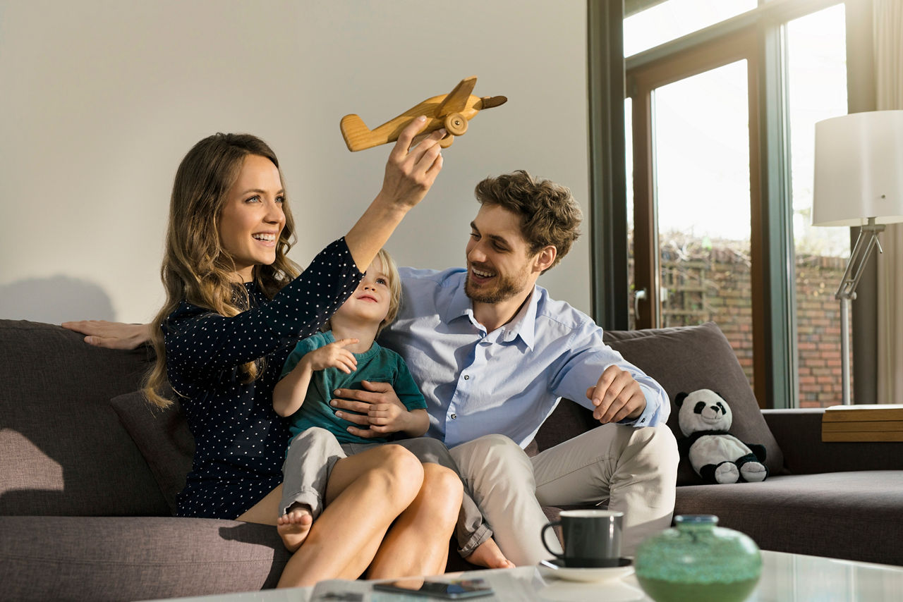 Germany, family portrait, parents and smiling toddler boy sitting on sofa in modern design living room and playing with wooden toy plane in front of large garden window