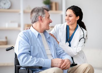 A healthcare professional smiles and talks to an older man sitting in a wheelchair. They are in a well-lit room with simple decor. The mood is friendly and supportive, focusing on the conversation.