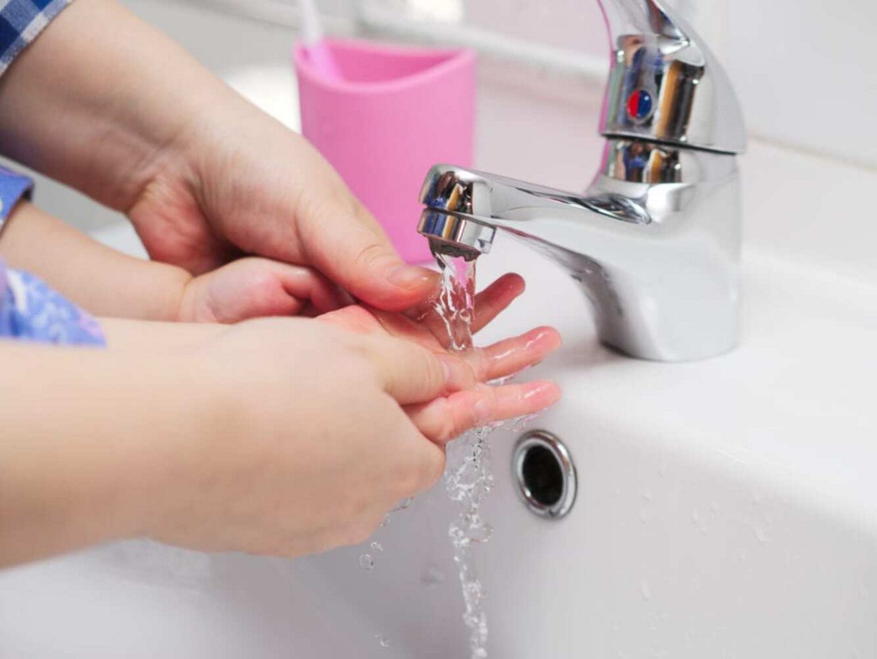 Un jeune enfant est guide par un adulte pour se laver les mains sous un robinet chrome brillant avec de l eau courante. Le decor semble etre un lavabo de salle de bain propre, avec un porte-brosse a dents rose visible en arriere-plan. L image met l accent sur l hygiene et les soins dans un environnement domestique.