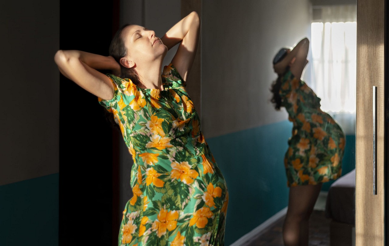 Une femme enceinte vetue d une robe a fleurs vertes et jaunes vibrantes pose a l interieur pres d un miroir. Le cadre se caracterise par une douce lumiere naturelle qui traverse une fenetre, creant une atmosphere chaleureuse et sereine. Le reflet dans le miroir ajoute de la profondeur a la composition, mettant en valeur le theme de la maternite.