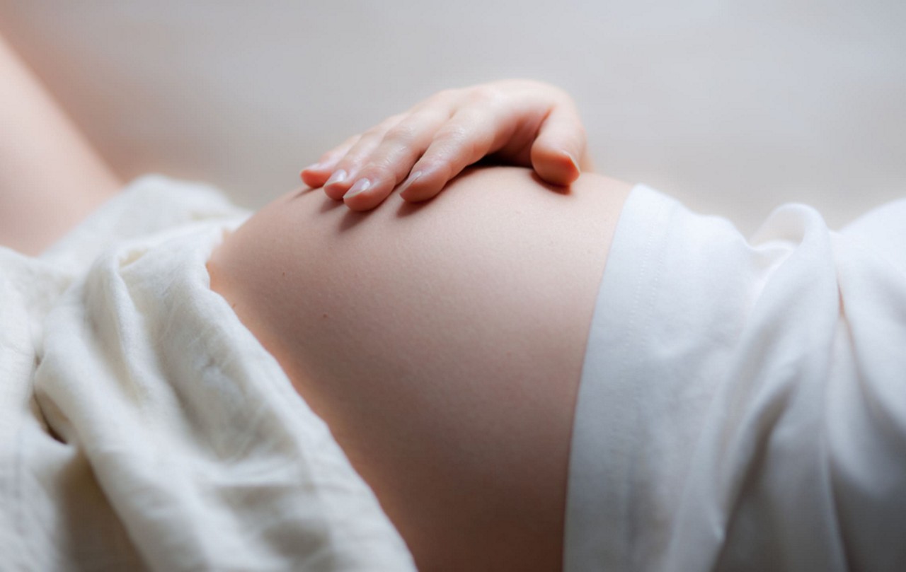 Gros plan d une femme enceinte posant doucement sa main sur son ventre. Le cadre est doux et serein, avec un eclairage naturel et un fond de tissu blanc. L image transmet une humeur calme et nourrissante, mettant l accent sur la maternite et l anticipation.