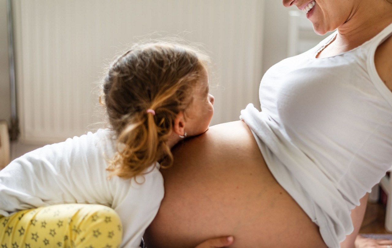 Un jeune enfant se penche pour embrasser le ventre d une femme enceinte, mettant en valeur un tendre moment de connexion. Le cadre semble etre a l interieur avec un eclairage naturel et un radiateur visible en arriere-plan. La femme porte un debardeur blanc et l enfant est vetu d une chemise blanche et d un pantalon jaune a motifs. L image transmet la chaleur, l amour et les liens familiaux.