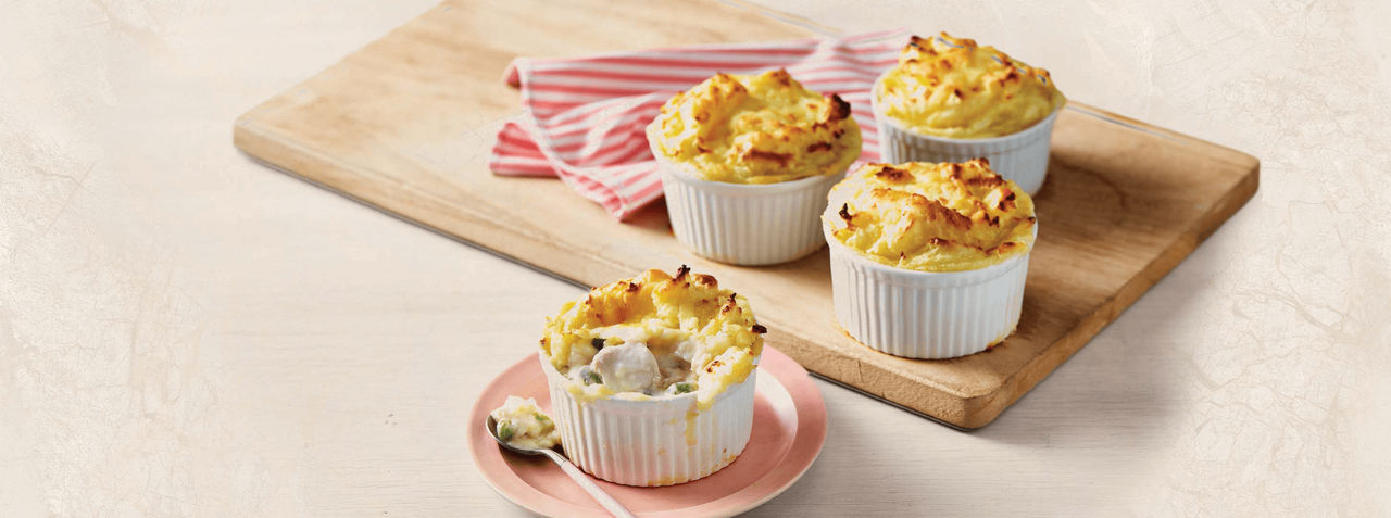 Four individual baked pot pies are presented in white ramekins, with golden-brown tops. Three are arranged on a wooden board with a red-striped cloth, while one is served on a pink plate with a spoon, revealing a creamy filling. The setting is bright and casual, emphasizing comfort food in a home kitchen environment.