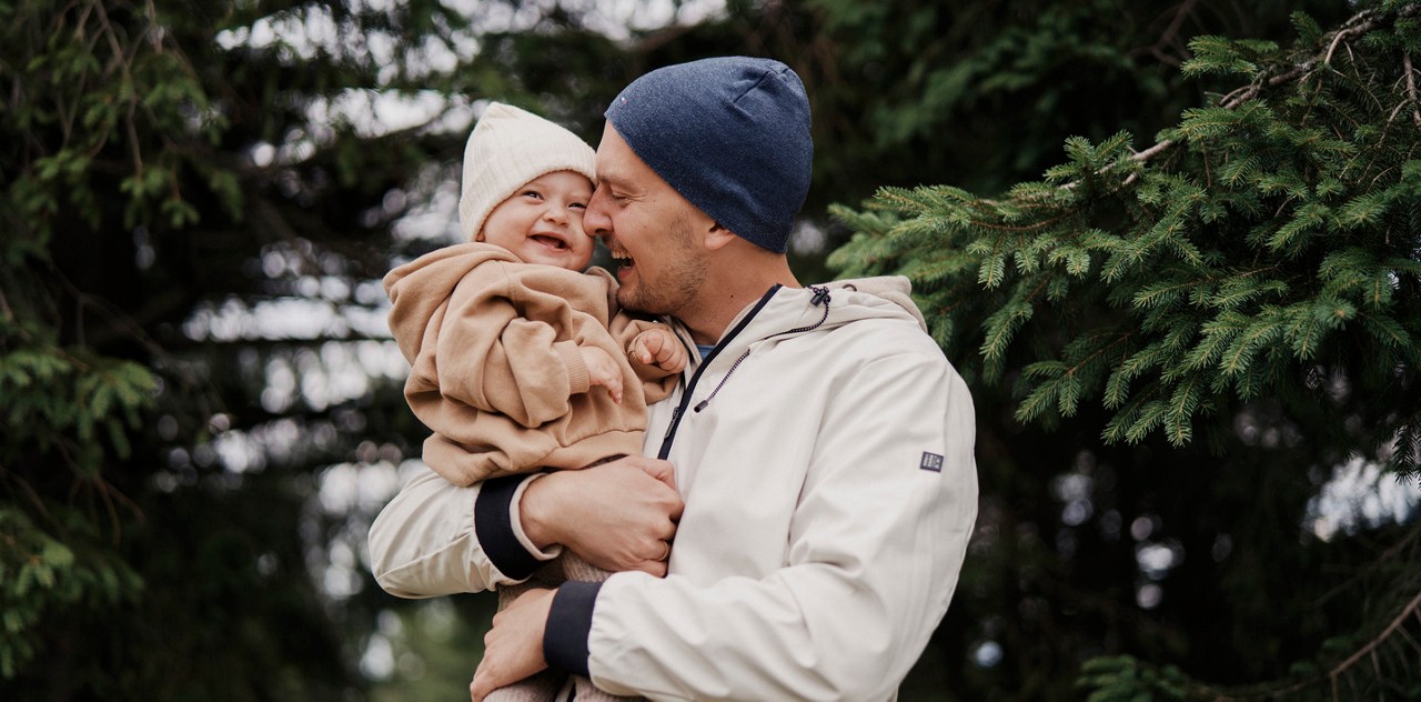 Un homme vetu d une veste blanche et d un bonnet bleu tient un bebe vetu d une tenue beige. La scene se deroule a l exterieur dans une foret luxuriante avec des arbres a feuilles persistantes en arriere-plan. L ambiance est sereine et naturelle, mettant l accent sur un lien avec la nature.