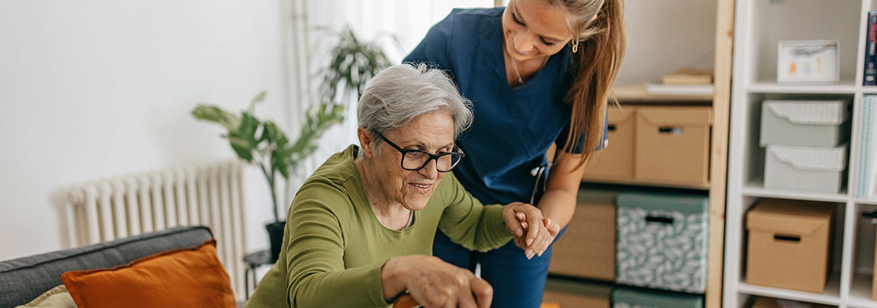 Female caregiver helping and supporting senior patient to stand up at home, senior women is sitting on sofa and holding walking cane