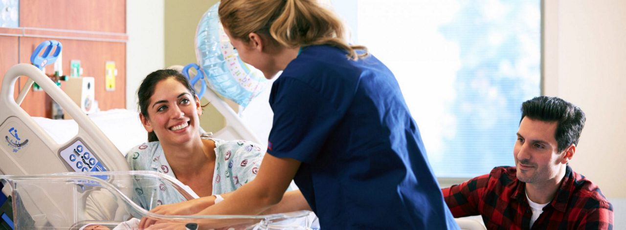 Eine Frau im Krankenhausbett lächelt eine Hebamme an, daneben sitzt ein Mann, im Vordergrund steht ein Babybett.