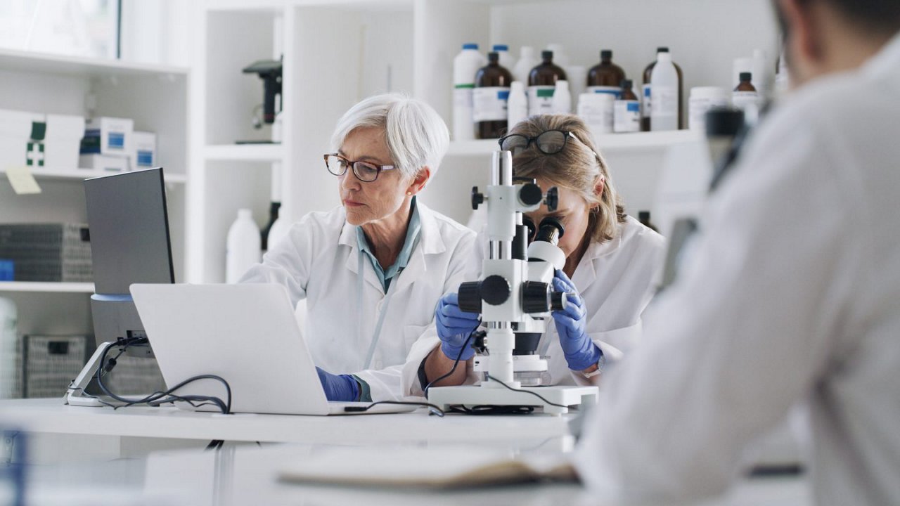 Zwei Forscherinnen im Labor arbeiten mit Mikroskop und Laptop, im Hintergrund stehen Laborflaschen und Regale.