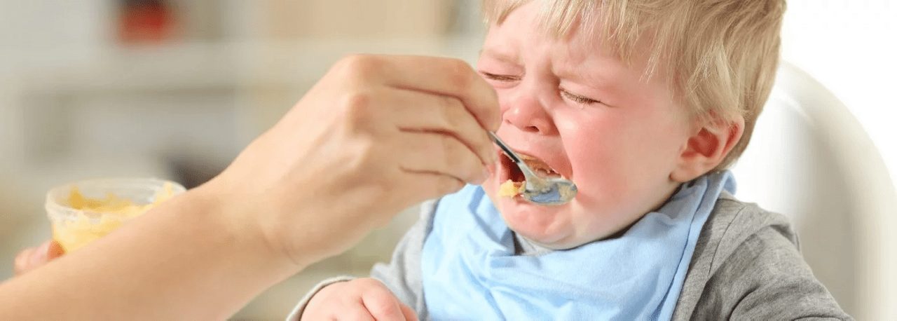 Ein weinendes Kleinkind wird mit einem Löffel Brei gefüttert, im Hintergrund ist ein heller Raum unscharf erkennbar.