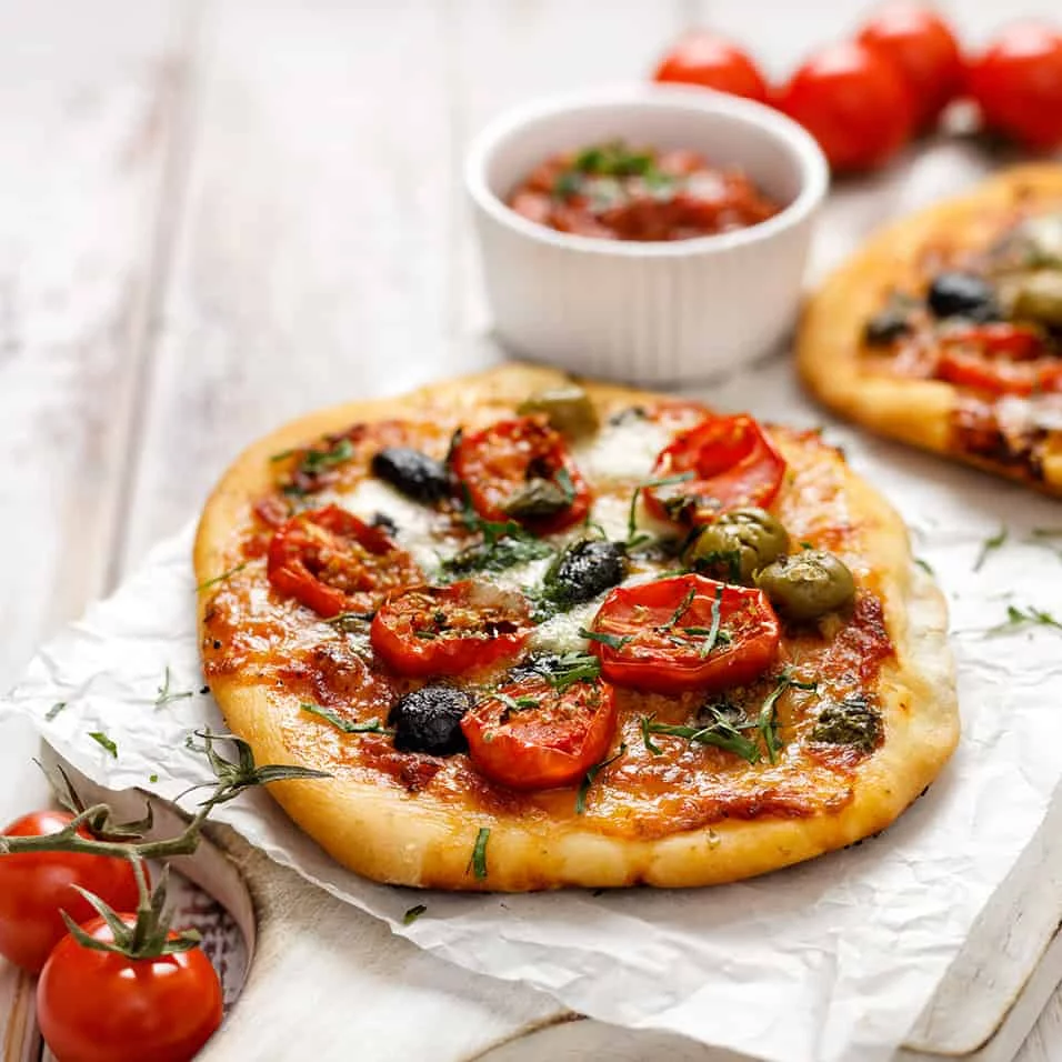 A close-up of a freshly baked pizza topped with vibrant cherry tomatoes, green and black olives, and melted cheese. The setting features a rustic wooden table with a bowl of tomato sauce and fresh tomatoes in the background. The image highlights the colorful ingredients and artisanal presentation.