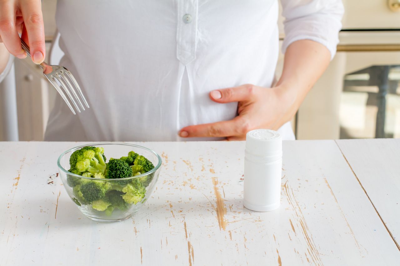 Pregnant female choosing between broccoli in glass bowl and vitamins, kitchen background