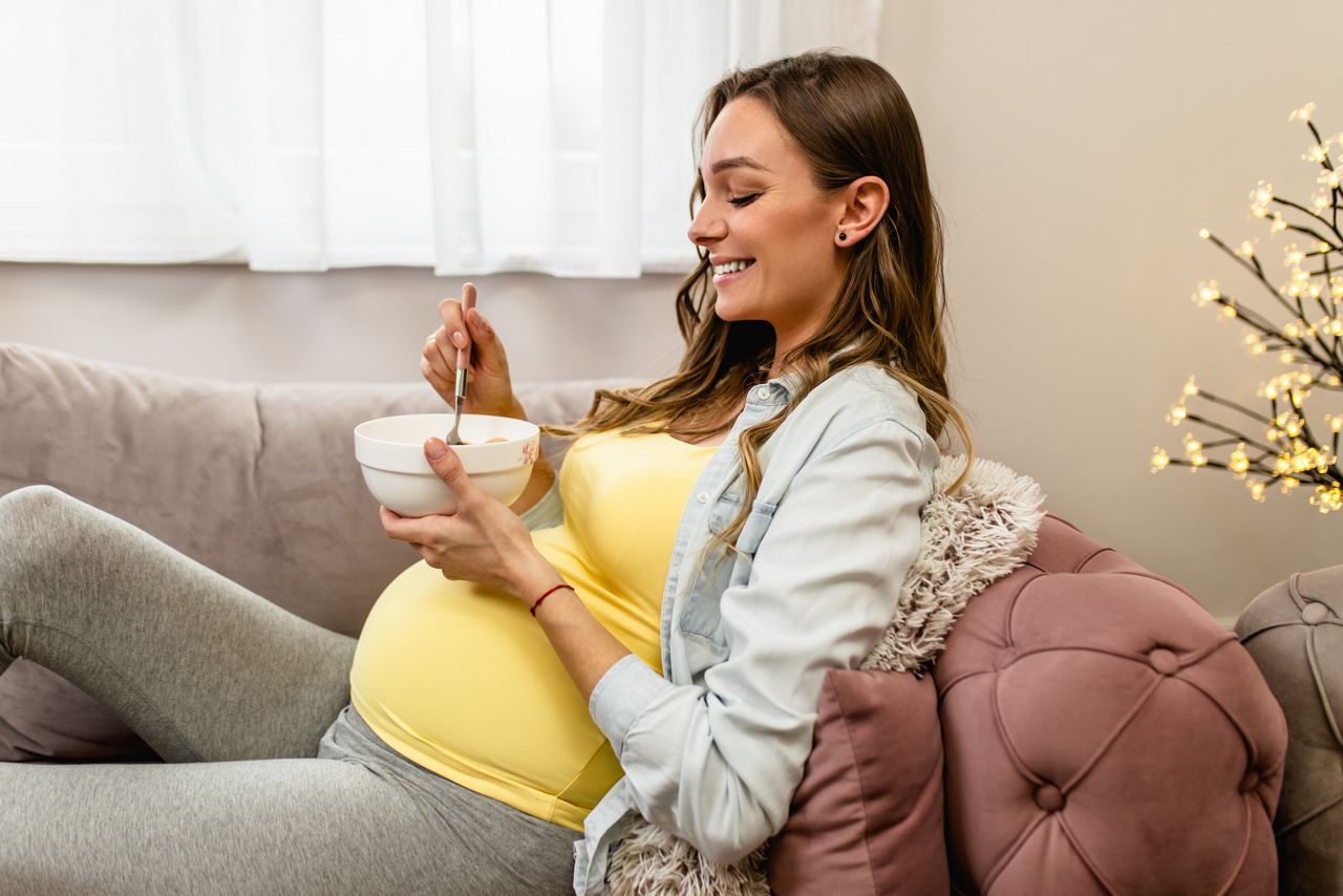 Beautiful young pregnant woman relaxing on sofa and eating corn flakes. Pregnancy concept.