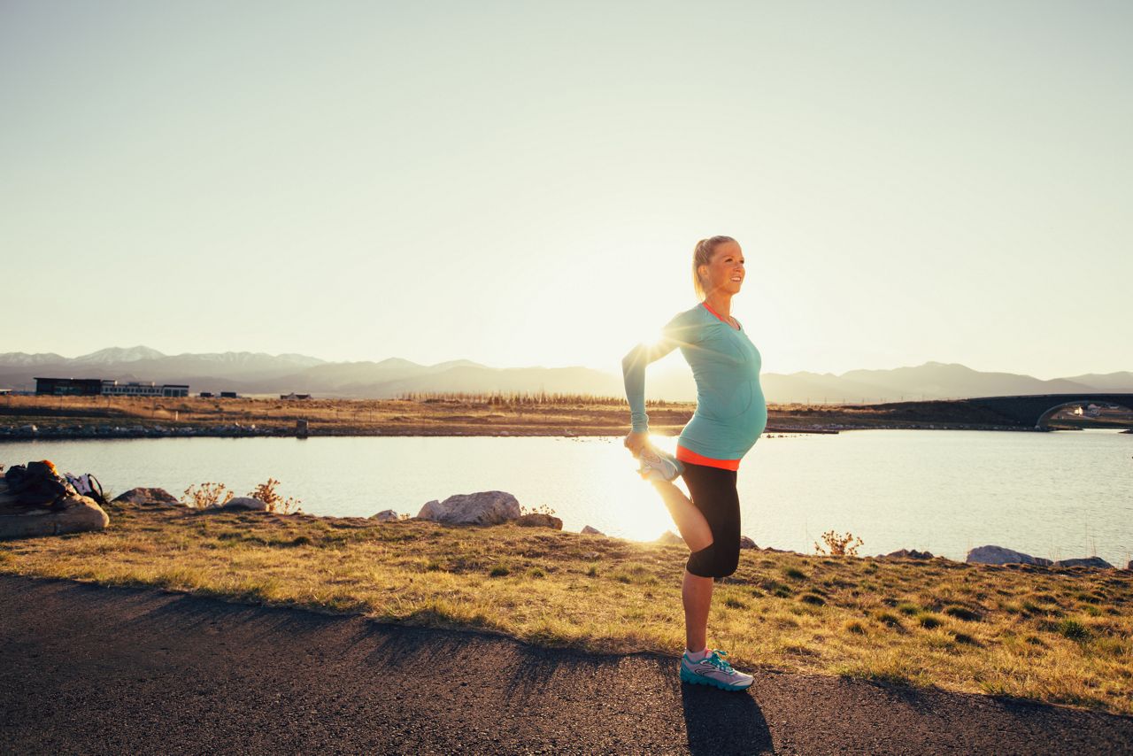 A pregnant female runner in a blue athletic shirt and running shoes is training while pregnant for her next race. She warms up by stretching with the sun flaring behind her. This woman enjoys running on trails and at sunset in the evening. Running is her favorite exercise to stay fit and happy.