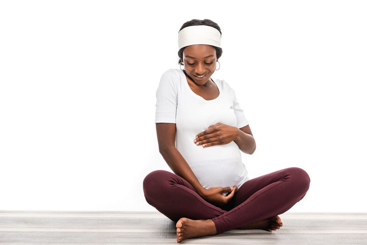 african american pregnant woman sitting on fitness mat and looking on belly isolated on white