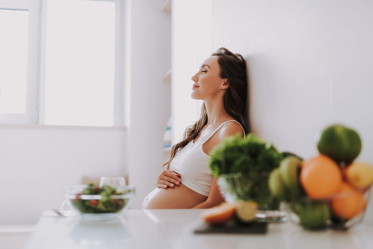 Profile side view of beautiful woman sitting on chair inside light room with closed eyes and holding hands on belly