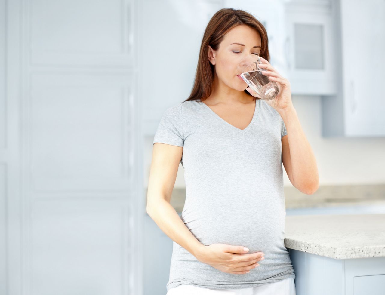 Young pregnant woman drinking a glass of water in her kitchen while holding her belly