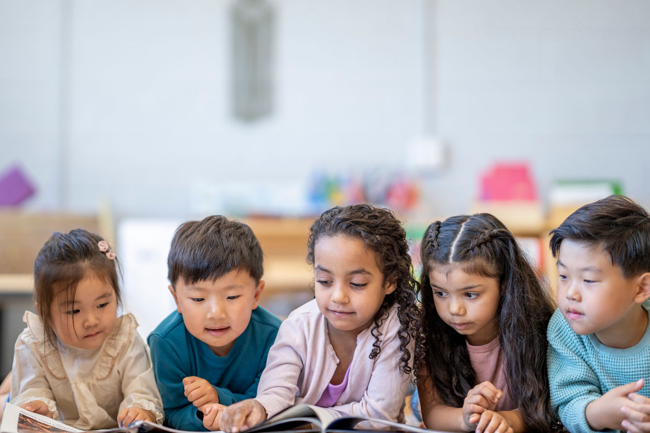 A small group of five Montessori children lay on their stomachs on the floor, side-by-side as they read together.  They ae each dressed casually and are focused on the stories.