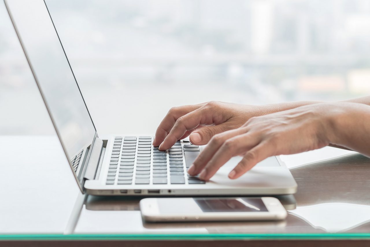 Hände tippen auf einem Laptop, daneben liegt ein Smartphone auf einem Glastisch vor verschwommenem Stadtpanorama.