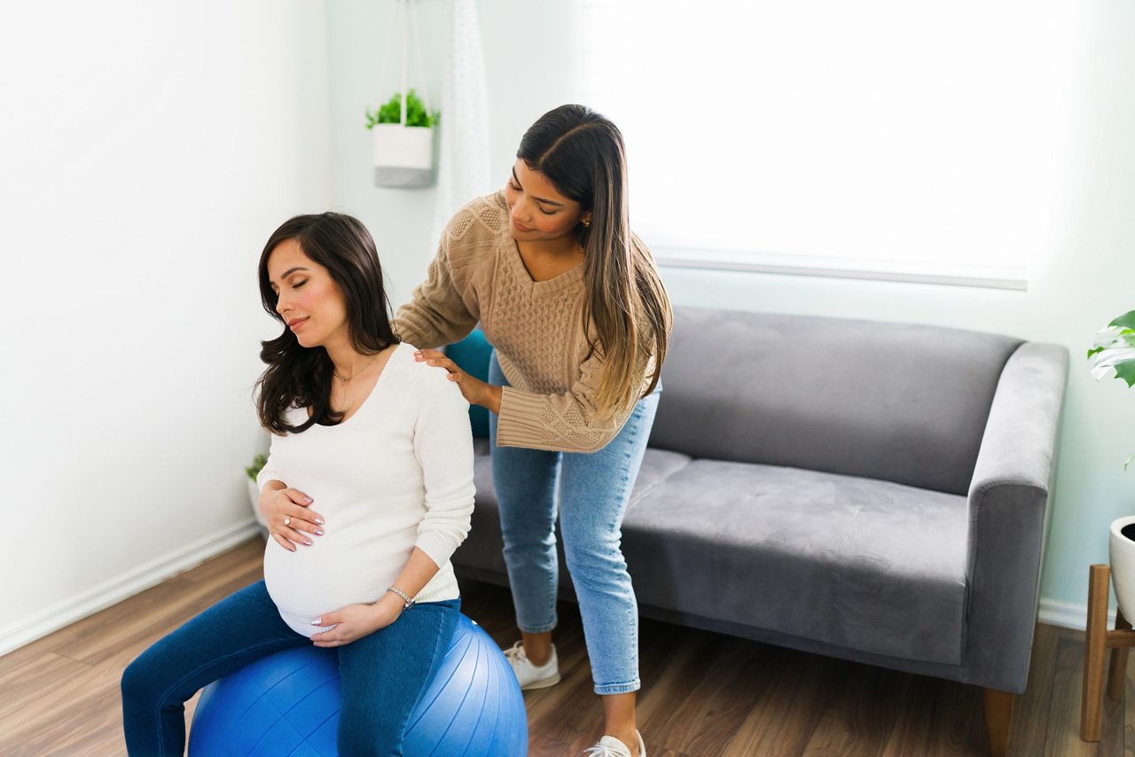 Attractive expectant mother sitting on a fitness ball in the living room and relaxing with a back massage from her lovely midwife getty images 1304285529