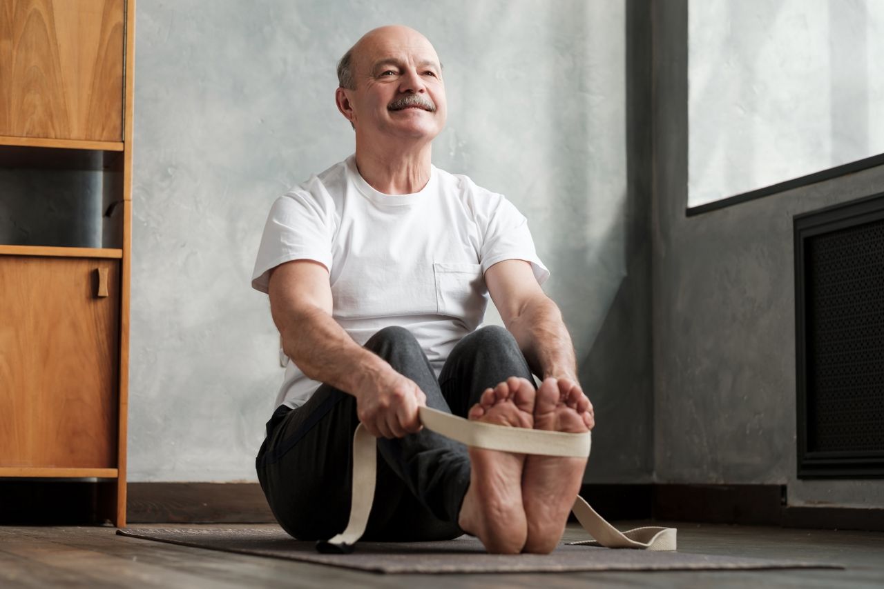 Senior hispanic man sitting in paschimottanasana or Intense Dorsal Stretch pose, seated forward bend posture, exercise for hips and spine at home using belt.