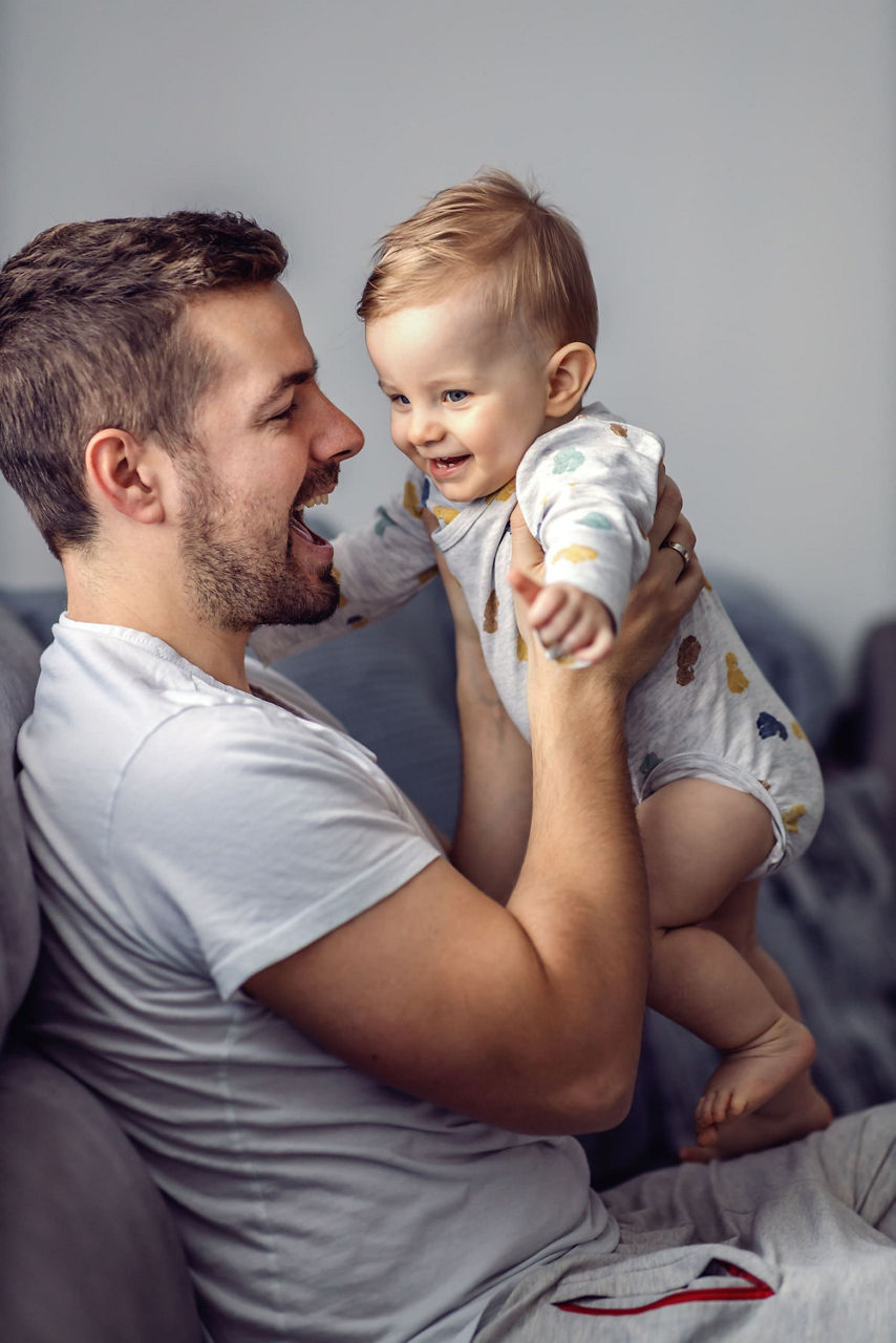 Un homme tient un bebe dans un environnement familial chaleureux et intime. Le bebe est vetu d une grenouillere blanche aux motifs colores, tandis que l homme porte un t-shirt blanc decontracte. Le cadre dispose d un eclairage tamise et d un canape aux tons neutres, creant une atmosphere calme et confortable.