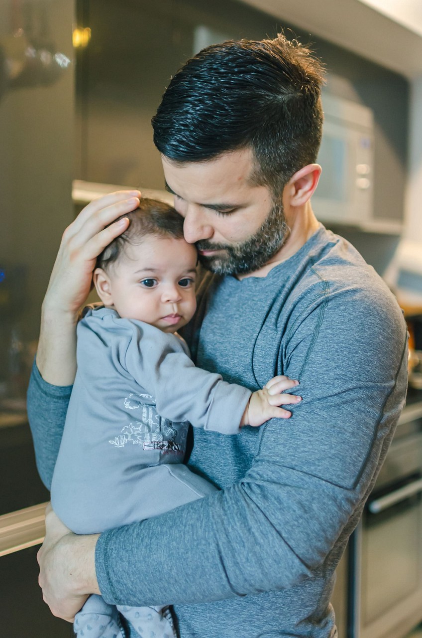 Un homme en chemise grise a manches longues tient un bebe vetu d une tenue grise. Le cadre est une cuisine moderne avec des appareils electromenagers elegants et une palette de couleurs neutres. La scene transmet la chaleur et les liens familiaux dans un environnement domestique contemporain.