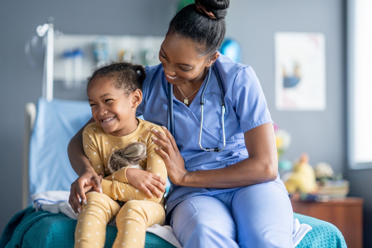 A sweet little pediatric patient, of African decent, sits on the end of his hospital bed as his nurse gives him a warm hug.  He is dressed comfortably in pajamas and giggling as he hugs his stuffed animal..