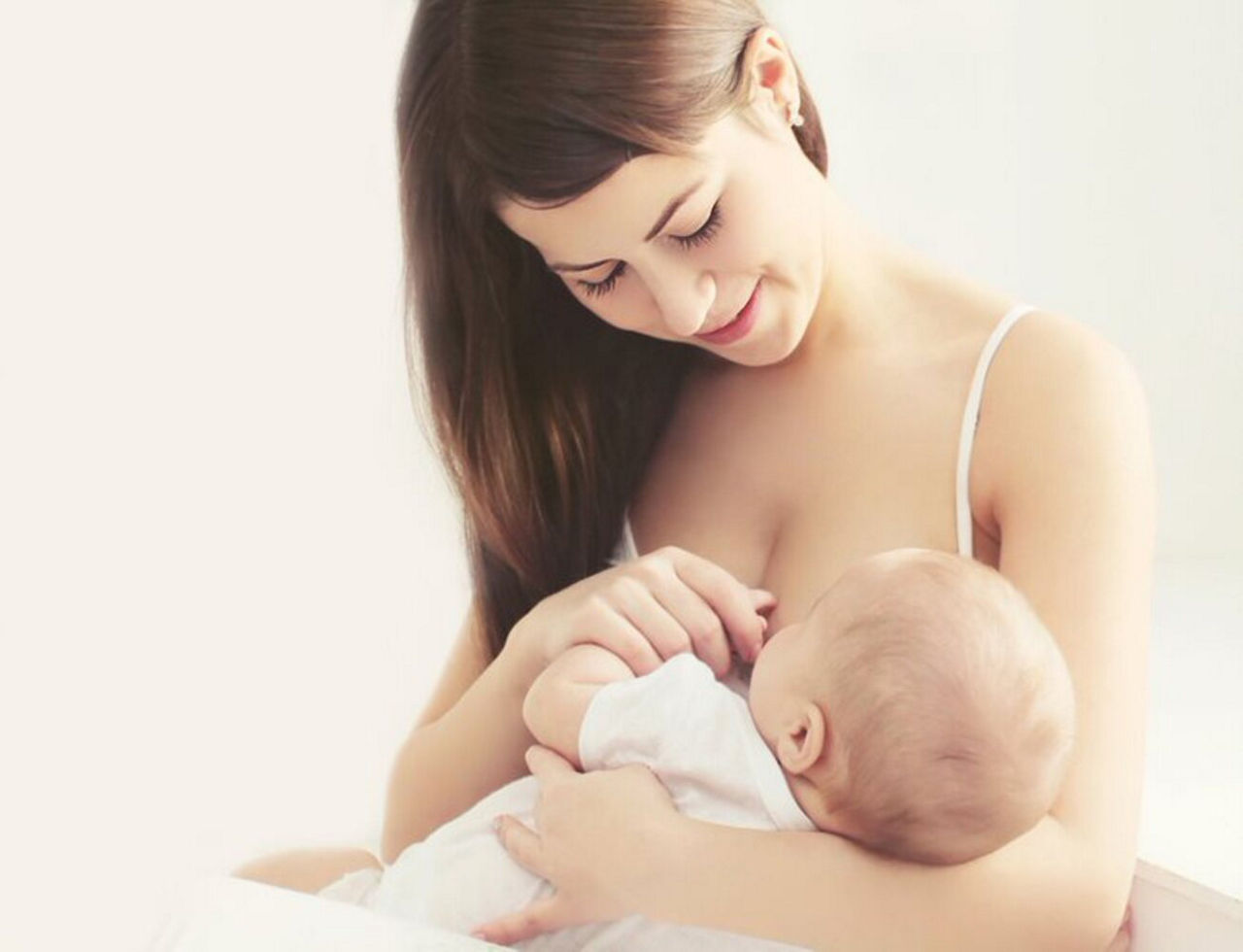 On voit une jeune mere allaiter son bebe dans un environnement interieur calme et minimaliste. Le cadre presente un eclairage doux et une palette de couleurs neutres, mettant l accent sur la chaleur et l intimite. Le bebe est vetu d une tenue blanche, completant l ambiance sereine generale de l image.