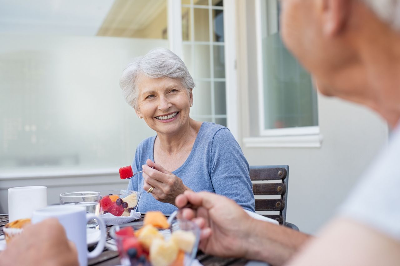 Happy senior woman eating fresh fruits during breakfast. Cheerful old lady with grey hair enjoying healthy breakfast with her husband outdoor. Elderly couple in conversation enjoying fresh fruit on the balcony.