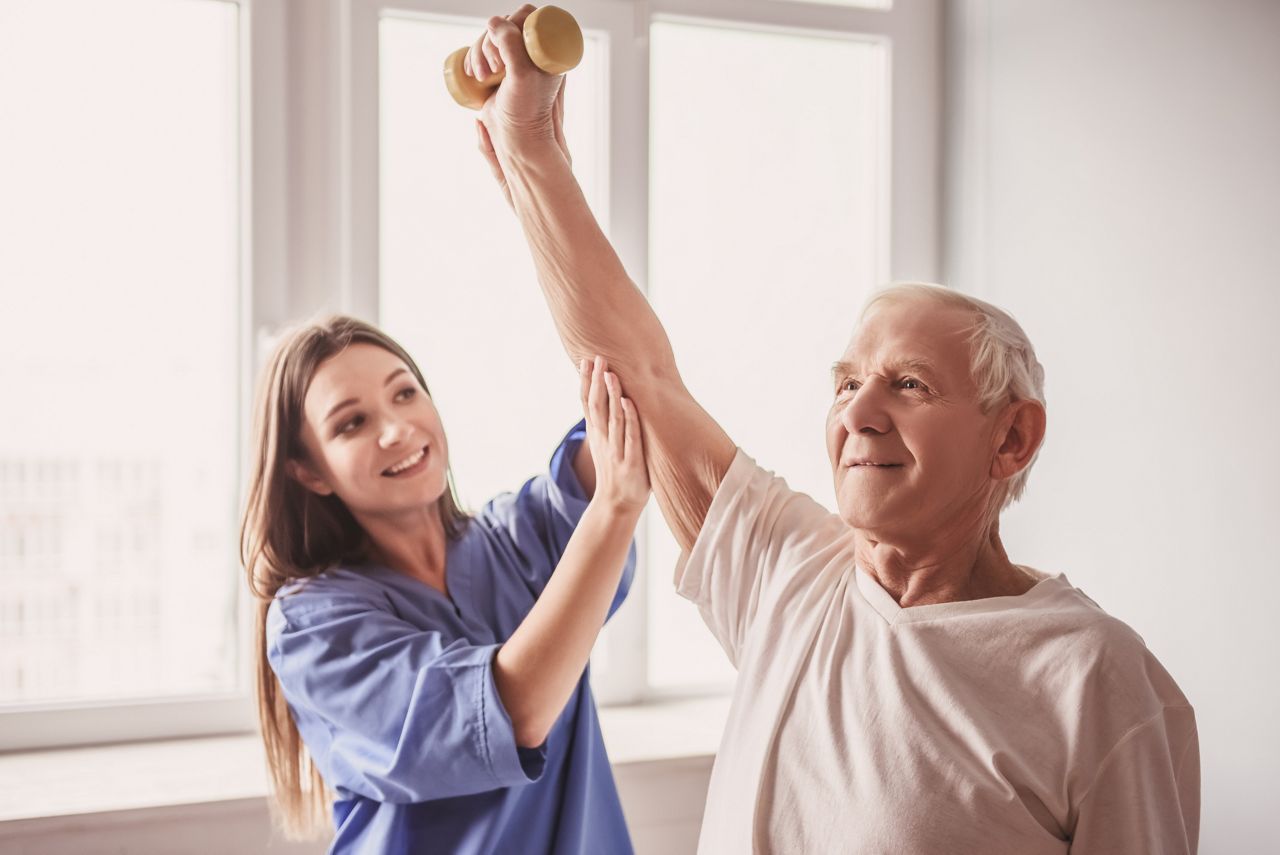 Handsome old man is doing exercises with dumbbells and smiling, in hospital ward. Attractive nurse is helping him